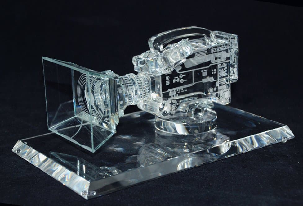 Clear video-camera miniature with projecting lens, front frame and top handle on a glass-like base