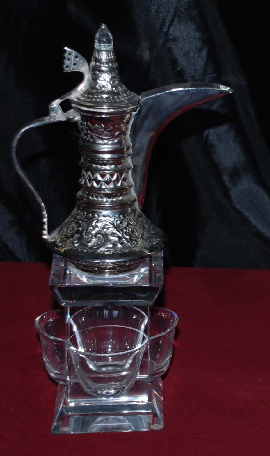 Decorative silver-toned Dallah with a long spout beside clear cups on a dark red surface